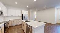 This single-floor unit showcases a contemporary design with a clean, neutral palette and high-quality finishes throughout. The open-concept kitchen serves as a central highlight, featuring a large island with quartz countertops, an undermount stainless steel sink, and a built-in dishwasher. Cabinetry combines crisp white upper cabinets with light wood-toned lower cabinets, accented by a classic white subway tile backsplash. Stainless steel appliances, including a French door refrigerator with ice and water dispenser, a range, and a microwave, complete the kitchen's functional appeal.

The living area is generously proportioned, featuring warm wood-look hard flooring, recessed lighting, and a ceiling fan. Large windows allow natural light to fill the space, offering views of the surrounding greenery. Bedrooms are carpeted for comfort, each with access to natural light through well-placed windows, and multiple walk-in closets equipped with wire shelving systems provide ample storage.

The bathroom features a soaking tub with full-height white grid tile surround, matte black hardware accents, and clean tile flooring. The primary bedroom is notably spacious with elevated ceilings and direct access to an en-suite bathroom with warm wood cabinetry.