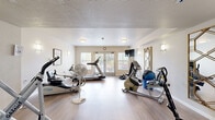 Located at 1585 Kipling St in Lakewood, Colorado, this dedicated exercise room offers a thoughtfully designed fitness space that balances functionality with a clean, welcoming aesthetic. The room features light hardwood-style flooring throughout, which contributes to a bright and open feel, complemented by soft white walls and warm sconce lighting positioned at regular intervals.

The space is well-equipped for a comprehensive workout, housing a treadmill, elliptical trainer, recumbent bike, rowing-style machine, weight bench, dumbbell rack, and a stability ball, among other accessories. A wall-mounted flat-screen television adds convenience for entertainment during exercise sessions.

Decorative gold-framed mirrors are mounted along the walls, serving both a practical and aesthetic purpose by enhancing the sense of space. A framed landscape painting adds a subtle artistic touch to the environment. Flush-mount ceiling lights provide consistent overhead illumination throughout the room.

Natural light enters through a set of large windows and glass-paneled French doors at one end of the room, which open to an outdoor area, allowing for ventilation and views of the surrounding landscape. The overall ambiance is clean, modern, and inviting, making this a functional and comfortable fitness facility.