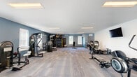 Located at 8681 Edison Plaza Dr in Fishers, Indiana, this fitness center offers a generously sized, single-floor exercise room designed to accommodate a wide range of workout routines. The space features a clean, modern aesthetic with a neutral color palette — crisp white ceilings contrast against cool blue-grey accent walls, creating an energizing yet focused atmosphere throughout.

The exercise room is outfitted with a comprehensive selection of equipment to support both cardio and strength training. Cardio options include treadmills, rowing machines, and stationary bikes, while the strength training area features multi-use cable machines, a functional training rig, weight benches, and a dumbbell rack. A wall-mounted flat-screen television adds an entertainment element to the space.

Durable, tile-patterned carpet flooring covers the entire floor, providing both comfort and practicality for varied exercise activities. Recessed flat-panel lighting fixtures illuminate the ceiling evenly, ensuring a well-lit environment. Multiple windows along the walls allow natural light to filter in, contributing to an open and inviting ambiance. The layout is thoughtfully organized to maximize usable floor space, with equipment distributed to allow for comfortable movement between stations. A dedicated entry door provides easy access to the facility.