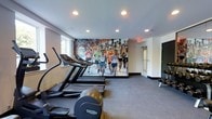 This fitness room offers a focused and motivating environment for cardiovascular training. The space is outfitted with a row of Technogym treadmills and an elliptical trainer, all positioned to face a striking, large-scale wall mural depicting a vibrant marathon race scene filled with runners in colorful athletic gear. This eye-catching artwork serves as a dynamic visual centerpiece, adding energy and inspiration to the workout experience.

The room features recessed ceiling lighting that provides bright, even illumination throughout, complemented by a window along one wall that introduces a degree of natural light. The flooring is covered in a durable grey carpet suited for a fitness environment. Along the far wall, a small selection of free weights is visible, offering additional options beyond the cardio equipment.

The layout is clean and functional, with enough open floor space to move comfortably between machines. An emergency exit sign is clearly visible, and a mirrored or glass-paneled door provides access. The overall design is modern and minimal, keeping the focus on the equipment and the bold mural that defines the room's motivating atmosphere.