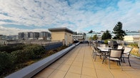 This rooftop terrace offers an expansive outdoor experience elevated above the surrounding cityscape. The deck is finished with large-format square paving tiles in a warm tan tone, providing a clean and durable surface across the entire space. Generous raised planter beds line the perimeter, filled with a variety of shrubs, ornamental grasses, and greenery that add a natural, landscaped quality to the urban setting. A dedicated outdoor dining area is furnished with multiple table-and-chair sets, making the terrace well-suited for gatherings and al fresco dining. Stairwell access structures with white lap siding are integrated into the layout, maintaining a tidy and functional design. Decorative ceramic planters with tropical-style plants are placed throughout, adding vibrant accents to the space. The terrace enjoys unobstructed 360-degree views of the surrounding urban environment, including nearby commercial buildings, residential towers, mature trees, and glimpses of a waterfront horizon. Under open skies, the space benefits from abundant natural light throughout the day, creating a bright and inviting atmosphere that makes this rooftop terrace a standout amenity.