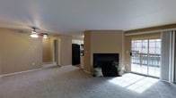 This single-floor residence features an open-concept layout that connects the main living area to the kitchen, creating a functional and cohesive living environment. The living room is finished with wall-to-wall carpeting and neutral beige walls, lending a warm, understated tone throughout. A black-surround fireplace serves as the room's focal point, positioned along a central wall and complemented by decorative accents nearby. A ceiling fan overhead provides practical climate control for the space.

Natural light enters generously through a sliding glass door on one side of the living room, which provides access to an outdoor area and offers views to the exterior. This door also contributes to the bright, airy feel of the main living space.

The kitchen is partially visible from the living area and features white cabinetry, a black refrigerator, and dark countertops, offering a functional workspace with a classic contrast of light and dark finishes. A hallway visible from the living room leads to additional areas of the unit, including what appears to be a closet and further rooms, suggesting a practical and well-organized floor plan throughout.