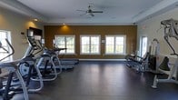 This single-floor fitness space is designed to provide a functional and comfortable workout environment. The room is anchored by warm, golden-yellow accent walls trimmed in white, creating an energizing yet inviting atmosphere. Three large white-framed windows line the main wall, allowing generous natural light to fill the space while offering views of the surrounding greenery outdoors.

The flooring consists of dark, durable material well-suited for fitness use, providing both stability and easy maintenance. A ceiling fan is centrally mounted overhead, ensuring proper air circulation throughout workouts. Recessed lighting supplements the natural light for consistent illumination at any time of day.

The cardio equipment area is prominently featured, with multiple treadmills and an upright exercise bike positioned along the windowed wall. Additional fitness equipment, including a rowing or multi-function machine, is stationed on the opposite side of the room. A wall-mounted television screen is also present, allowing users to stay entertained during exercise sessions. Equipment racks along the walls offer organized storage for accessories, contributing to the room's clean and orderly layout.