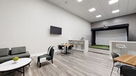 Located at 8850 River Bend Pkwy in Indianapolis, IN, this indoor golf simulator suite offers a well-appointed, single-floor space designed for both recreation and entertainment. The room is finished with light gray luxury vinyl plank flooring, crisp white walls, and a drop ceiling with recessed panel lighting that creates a bright, clean atmosphere throughout.

At the heart of the space is a TruGolf simulator bay, complete with an artificial turf hitting mat and a projection screen enclosure. A dedicated computer workstation sits adjacent to the bay for managing simulator settings, and a track-mounted spotlight illuminates the hitting area. A wood golf club rack provides convenient equipment storage nearby.

The social area features a central white bar-height counter flanked by bar stools, along with multiple bistro-style tables and chairs for guests. A wall-mounted flat-screen TV anchors one end of the room, while a comfortable sectional sofa and oval coffee table create a relaxed lounge area. Decorative wall art adds a subtle design touch.

A built-in white cabinetry unit with a beverage refrigerator and open display shelves serves as a refreshment station, with a doorway leading to a restroom. The space is thoughtfully laid out to comfortably accommodate players and spectators alike, making it a versatile venue for casual rounds, group outings, or golf instruction.