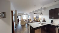 This single-floor unit offers a thoughtfully designed open-concept layout that integrates the kitchen, dining, and living spaces into one cohesive environment. The kitchen serves as a central focal point, featuring dark espresso cabinetry, granite countertops, a stainless steel sink, and a built-in dishwasher. Two brushed nickel pendant lights hang above the kitchen counter, providing both task lighting and a stylish design element.

The open layout flows naturally into the dining and living areas, where rich, dark hardwood-style flooring runs throughout, adding warmth and continuity to the space. The living area is generously sized, accommodating comfortable seating arrangements, and is brightened by natural light streaming through large windows dressed with sheer curtains. A ceiling fan is centrally positioned in the living area, enhancing airflow and comfort.

The neutral wall tones and white ceilings contribute to an airy, clean aesthetic throughout. The overall ambiance is modern yet inviting, with a balance of functional design and contemporary finishes that make the space both practical and visually appealing.