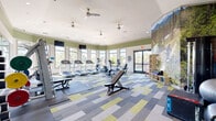 Located at 4505 Dublin Blvd in Colorado Springs, CO, this fitness center offers a generously sized, single-floor workout facility designed to support a full range of exercise routines. The space features a bright, open layout illuminated by large windows along multiple walls, providing ample natural light and views of the surrounding landscape. Ceiling fans are installed throughout to maintain comfortable airflow during workouts.

The cardio zone is well-appointed with multiple treadmills, elliptical trainers, and stationary bikes, each positioned to take advantage of the natural light and outdoor views. Wall-mounted flat-screen televisions provide entertainment during cardio sessions. The strength training area includes a cable machine, a squat rack loaded with bumper plates, an adjustable weight bench, and a full dumbbell rack offering a wide range of weights.

A striking Colorado mountain landscape mural serves as a dramatic focal point on a curved accent wall, complementing the sage green upper walls and white ceilings. The floors are covered in a contemporary patchwork-style carpet in shades of gray, blue, and yellow-green, adding a modern, energetic feel to the space. Additional equipment such as kettlebells, medicine balls, and stability balls round out the functional training options, making this facility a comprehensive resource for residents.