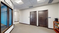 The Clubhouse Lobby at Falcon Heights in Rochester, Minnesota, serves as the welcoming gateway to the community's amenities. The lobby features a clean, modern design with neutral-toned carpet, crisp white walls, and a drop ceiling with recessed panel lighting that creates a bright, well-lit environment. A glass entry door with full-height sidelights draws in natural light from the exterior, while a welcoming seating area with a cushioned accent chair, a dark media console, a decorative electric fireplace, and tasteful artwork provides a comfortable first impression for residents and guests alike. A small shelving unit near the entrance offers a convenient coffee station, adding a hospitable touch to the space. Two dark-stained, solid wood interior doors with panel detailing lead to private office spaces, their rich tones complementing the warm wood base trim throughout. A standout feature of the lobby is a large interior window framed in matching dark wood trim, offering a clear view into the community's fitness center — branded as "The Nest" — which is visible with a full array of exercise equipment. The overall ambiance is polished and functional, balancing a professional aesthetic with an inviting, community-oriented atmosphere.