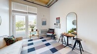 Located at 7141 Woodbine Ave in Sacramento, CA, this two-story loft offers a thoughtfully designed layout spread across two levels. The main floor serves as the heart of the space, featuring an open-plan living area with impressively tall ceilings accented by warm wood paneling and exposed ductwork, lending an industrial-chic aesthetic. Large French doors flood the living area with natural light and provide views of the surrounding greenery, creating an airy, inviting ambiance. The kitchen is efficiently designed with light wood cabinetry, a clean white countertop, built-in oven, cooktop, and a full-size refrigerator — maximizing functionality without sacrificing style. Two full bathrooms serve the unit, each featuring clean, modern finishes including a tiled shower-tub combo, LED-lit mirror, and wood-accented storage cabinetry. A ladder leads to the upper loft level, which offers a private, cozy retreat ideal for use as a sleeping area or additional lounge space. Throughout the space, light-toned flooring and white walls provide a neutral canvas that enhances the sense of openness. This loft combines smart, space-efficient design with modern industrial character in a well-located Sacramento address.