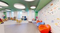 This thoughtfully designed children's play space offers a bright, engaging environment spread across a single open floor. The interior is defined by a distinctive teal and green ceiling accented with large, circular pendant light fixtures that provide warm, even illumination throughout. Tall windows along the perimeter flood the space with abundant natural light while offering views of the surrounding cityscape, creating an open and airy atmosphere.

The layout is organized into several dedicated activity zones. A creative play area features child-sized tables and chairs alongside shelving units stocked with toys, dollhouses, and building sets. A reading nook with built-in wooden bookshelves houses an accessible collection of books for young readers. A soft play zone is equipped with interlocking foam floor mats and cushioned floor pillows, ideal for free play and relaxation. A large whiteboard wall provides an additional interactive element for creative expression.

A standout feature is the expansive illustrated world map mural, rendered in a playful, animal-themed style that serves as both a decorative focal point and an educational element. Complementing the mural are a row of bold, molded pod chairs in orange and blue, adding a fun, modern design touch. Throughout the space, vibrant green accent walls, colorful furnishings, and durable flooring reinforce the energetic yet welcoming character of this purpose-built children's environment.