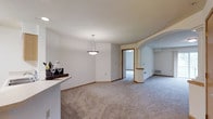 This single-floor residence offers a thoughtfully designed open-concept layout that promotes a sense of spaciousness and ease of movement throughout. The main living area is finished with soft, plush wall-to-wall carpeting in a neutral tone, complemented by clean white walls and warm wood-trimmed doorways that add a touch of classic elegance. A stylish pendant light fixture serves as a focal point, illuminating the transition between the dining and living spaces. A partial-height counter or bar area provides a natural division between rooms while maintaining visual openness.

A graceful arched opening with a decorative column detail adds architectural character, leading into an additional living or flex space that offers versatility for various uses. Doorways provide access to further rooms, hinting at a well-considered floor plan designed for comfortable single-level living.

Beyond the interior, the outdoor surroundings are truly a standout feature. The property enjoys views of a beautifully maintained landscape, including a serene pond with a natural stone waterfall feature, rolling green lawns, and a backdrop of mature evergreen and deciduous trees. This peaceful natural setting enhances the overall ambiance of the space, making it an ideal retreat for those who appreciate tranquil, scenic surroundings.