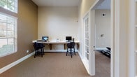 The business center is a thoughtfully designed single-floor workspace finished with warm neutral tones and carpeted flooring throughout. The room is anchored by a long, wall-mounted white desk that spans one side of the space, accommodating multiple workstations equipped with desktop computers and a printer, each paired with cushioned bar-height chairs. The walls are painted in a two-tone scheme of soft beige and warm taupe, accented by clean white baseboards and trim. A large window fitted with horizontal blinds allows natural light to fill the room, offering views of the surrounding greenery and outdoor environment. An arched transom window above adds architectural character while drawing in additional daylight. Decorative artwork in cool aqua tones adorns the wall, providing a calming visual element. Built-in panel access points are neatly integrated into the walls. A set of white-framed French doors with glass panes connects the business center to an adjacent fitness facility, creating a seamless flow between amenity spaces. The overall atmosphere is clean, functional, and professional.