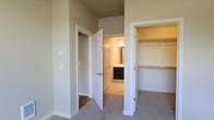 This single-floor space showcases a thoughtfully designed bedroom area that prioritizes functionality and convenience. The room is finished with neutral beige walls and plush carpeting, creating a calm and comfortable atmosphere. Notably tall walls extend upward, lending an open, airy feel to the space.

A white-paneled door opens directly into a private ensuite bathroom, which features warm lighting, a large mirror, and dark-toned cabinetry beneath the vanity, providing ample storage. The bathroom's light-toned flooring contrasts tastefully with the darker cabinet finish.

Adjacent to the bathroom entrance is a generously sized reach-in closet, equipped with built-in double hanging rods and a shelf above, offering well-organized storage for clothing and accessories. The closet is finished with the same neutral tones found throughout the room, maintaining visual consistency.

Clean white trim and door casings frame each entryway, providing a crisp contrast to the warm wall tones. The overall design emphasizes practicality while maintaining a cohesive, understated aesthetic throughout the space.
