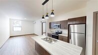This well-designed 1-bedroom, 1-bathroom unit offers a thoughtfully arranged single-floor layout that maximizes functionality and comfort. The open-concept kitchen and living area serves as the heart of the unit, anchored by a central island with a light stone-look countertop, double sink, and a trio of stylish pendant lights overhead. The kitchen features rich dark brown cabinetry, stainless steel appliances including a refrigerator, electric range, and microwave, all set against clean light countertops. Wood-tone laminate flooring flows consistently throughout the space, adding warmth to the bright, neutral palette. The living area is open and airy, benefiting from a wall-mounted mini-split HVAC system for efficient climate control. The bedroom is a comfortable, well-proportioned room bathed in natural light from a large window with outdoor views of lush, snow-dusted evergreens, creating a tranquil atmosphere. The bathroom is generously sized and includes a wood-toned vanity with a stone-look countertop, a walk-in shower, and convenient in-unit laundry access. A dedicated closet provides ample storage. Clean lines, neutral tones, and modern finishes define the overall ambiance of this practical and contemporary space.