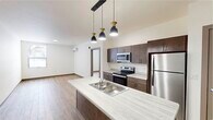 This well-designed 1-bedroom, 1-bathroom unit offers a thoughtfully arranged single-floor layout that maximizes functionality and comfort. The open-concept kitchen and living area serves as the heart of the unit, anchored by a central island with a light stone-look countertop, double sink, and a trio of stylish pendant lights overhead. The kitchen features rich dark brown cabinetry, stainless steel appliances including a refrigerator, electric range, and microwave, all set against clean light countertops. Wood-tone laminate flooring flows consistently throughout the space, adding warmth to the bright, neutral palette. The living area is open and airy, benefiting from a wall-mounted mini-split HVAC system for efficient climate control. The bedroom is a comfortable, well-proportioned room bathed in natural light from a large window with outdoor views of lush, snow-dusted evergreens, creating a tranquil atmosphere. The bathroom is generously sized and includes a wood-toned vanity with a stone-look countertop, a walk-in shower, and convenient in-unit laundry access. A dedicated closet provides ample storage. Clean lines, neutral tones, and modern finishes define the overall ambiance of this practical and contemporary space.