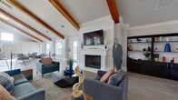 This expansive single-floor gathering space showcases a striking architectural design centered around soaring vaulted ceilings with rich, exposed wood beams that run the length of the open interior. Light hardwood flooring flows throughout, complementing the crisp white walls adorned with classic wainscoting and crown molding that lend a refined, traditional character to the space.

The primary lounge area features a white-surround fireplace with a wall-mounted television above, flanked by a dark built-in bookcase and a large arched window that frames outdoor views. Multiple seating arrangements — including plush sofas, leather armchairs, and accent chairs in navy and brown — create inviting conversational zones throughout. A geometric pendant light fixture adds a contemporary touch to the open central area.

An intimate nook features a round marble-top table with tufted bench seating beneath a curated gallery wall, bathed in natural light from clerestory windows above. The space also includes a bar-height counter area and a casual dining arrangement, making it well-suited for both social gatherings and quiet meetings. Tall windows and glass-panel doors along multiple walls flood the interior with natural light and offer views of the surrounding landscape, enhancing the bright, welcoming ambiance throughout.