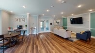 Located at 4826 Van Buren Blvd in Riverside, California, this single-floor community clubhouse is designed to serve as a versatile and inviting gathering space. The interior is finished with rich hardwood-style flooring that runs throughout, complemented by soft blue-gray walls, crisp white crown molding, and decorative column accents that lend the space a polished, cohesive aesthetic.

The open layout is divided into several functional zones. The lounge area features a contemporary white sofa, a wall-mounted flat-screen television, and curated wall art, creating a comfortable setting for relaxation or socializing. Adjacent to the lounge is a dedicated workstation area outfitted with a built-in desk, computer monitor, and a stainless steel dual water fountain — ideal for residents needing a quiet space to work or connect.

A separate dining nook accommodates a round table with seating for multiple guests, set alongside a rustic sideboard and tropical greenery that adds warmth to the corner. The kitchenette is well-appointed with white shaker-style cabinetry, a striking blue mosaic tile backsplash, stainless steel appliances including a dishwasher and refrigerator, a microwave, and a double sink. A window in this area allows natural light to filter in. Recessed lighting throughout ensures the entire space is bright and welcoming at all hours.