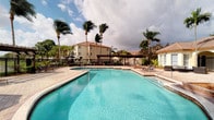 This outdoor amenity space embodies the essence of Florida resort-style living, centered around a generously sized, crystal-clear swimming pool with a classic curvilinear design. The pool deck is finished in light-toned pavers, providing ample space for sunbathing and leisure. Surrounding the pool, tall palm trees sway in the breeze, lending a tropical atmosphere to the area. A structured pergola runs along the back of the pool deck, offering shaded seating and a row of lounge chairs for relaxation. Adjacent to the pool, a well-appointed patio area features durable outdoor furniture, including a round dining table with chairs gathered beneath a large navy umbrella — ideal for al fresco gatherings. This patio sits alongside a building with a Mediterranean-influenced roofline featuring terracotta tiles, arched windows, and warm stucco exterior walls framed by lush, manicured landscaping. Tropical plantings and greenery border the space, contributing to a private, resort-like feel. The overall ambiance is vibrant and inviting, designed for outdoor recreation and relaxation in a warm, sun-drenched setting.