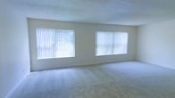 This single-floor space features a spacious, open living area that serves as the central focus of the layout. The room is finished with wall-to-wall carpeting in a neutral beige tone, providing a soft, comfortable foundation underfoot. Two generously sized windows fitted with vertical blinds line the exterior wall, allowing abundant natural light to flood the interior and creating a bright, airy atmosphere throughout the day. The walls are painted in a clean, light neutral shade that complements the soft flooring and enhances the sense of openness. A textured ceiling adds subtle architectural character to the room. The minimalist design of the space lends itself well to a variety of furniture arrangements and décor styles, making it highly adaptable to personal preferences. The overall ambiance is calm and understated, with the natural light serving as a primary design element that keeps the space feeling fresh and welcoming. The combination of neutral finishes, ample window coverage, and open layout makes this a functional and versatile living environment suited to a range of uses.