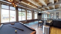 The Lofts at Helmetta offers a thoughtfully designed amenity lounge that blends industrial aesthetics with inviting comfort. The space features soaring ceilings with exposed wood planking, visible ductwork, and Edison bulb pendant lighting that together create a warm yet edgy loft-style atmosphere. Large, oversized windows line the walls, flooding the interior with abundant natural light and offering views of the surrounding outdoor scenery.

The game area is a central highlight, anchored by a full-size billiards table with a slate-blue felt surface and a classic shuffleboard table positioned along the windowed wall. Hardwood flooring transitions seamlessly into patterned teal carpeting that defines distinct zones within the space. A wet bar with dark cabinetry and granite countertops provides a social focal point, complemented by high-top bar seating ideal for casual gatherings.

A dedicated lounge and seating area features a round wooden table surrounded by cushioned chairs with striped upholstery, perfect for card games or conversation. Framed decorative artwork adorns the walls, adding personality to the space. Mounted flat-screen televisions enhance the entertainment offering. The combination of thoughtful design details, versatile seating arrangements, and abundant amenities makes this communal lounge a standout feature of The Lofts at Helmetta.