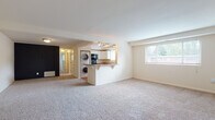 Located at 8001 York Rd in Towson, Maryland, this single-floor residence offers a practical and functional layout designed for comfortable everyday living. The expansive main living area is finished with plush wall-to-wall carpeting and neutral white walls, creating a bright and open atmosphere. A large window allows natural light to pour in while offering views of the surrounding greenery. The kitchen is positioned at the heart of the home, defined by a convenient peninsula with a breakfast bar overhang. It features white cabinetry, dark laminate countertops, a stainless steel sink positioned beneath a window, and a black dishwasher. A stacked washer and dryer unit is neatly integrated into the kitchen space, providing the convenience of in-unit laundry. The bedroom areas are generously proportioned, with soft carpeting underfoot and an impressive amount of built-in storage, including dual closets with shelving and hanging rods. The bathroom includes wood-look flooring, a sliding glass shower enclosure, and a window for natural ventilation. An electrical panel is accessible within the living space, and the unit benefits from a dedicated entryway. Throughout, the clean, neutral palette offers a versatile canvas well-suited to a variety of furnishing styles.
