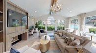 This thoughtfully designed single-story space combines modern elegance with functional comfort across several distinct areas. The main lounge is anchored by soaring vaulted ceilings and a striking brass chandelier, creating a grand yet inviting atmosphere. A large arched window floods the room with natural light and frames lush outdoor greenery, while recessed lighting complements the airy aesthetic. The lounge features a built-in entertainment center with a fireplace, open shelving, and an integrated workstation with dual monitors—ideal for both relaxation and productivity.

Adjacent to the lounge, a sunlit dining and lounge corridor runs along a wall of floor-to-ceiling sliding glass doors and elegant patterned drapes, offering direct views and access to a well-maintained outdoor patio. The light tile flooring continues seamlessly throughout, enhancing the cohesive, open feel of the space.

The kitchenette is efficiently appointed with cream cabinetry, a stainless steel refrigerator, a built-in microwave, open display shelving with gold-framed accents, and a peninsula with seating. Pendant lighting with a warm gold finish adds a refined touch. The overall palette of warm neutrals, soft blues, and gold accents creates a polished, welcoming environment throughout.