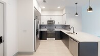 This single-floor space showcases a thoughtfully designed interior with a clean, modern aesthetic. The kitchen is a standout feature, outfitted with two-tone cabinetry — crisp white upper cabinets contrasting with dark lower cabinets — paired with stainless steel appliances including a gas range, built-in microwave, refrigerator, and dishwasher. A patterned geometric tile backsplash adds visual interest, while light-toned flooring and recessed lighting complete the contemporary look. A pendant light fixture accents the space with an industrial touch.

The living area is open and airy, benefiting from natural light streaming through a large window that offers views of the surrounding building exterior. Light hardwood-style flooring, crisp white walls, and a ceiling fan contribute to the bright and welcoming atmosphere.

The bedroom continues the clean design language with soft carpeting, a ceiling fan, and a generously sized window that draws in natural light and frames exterior views, creating an inviting and restful retreat.

The bathroom is well-finished with wood-look tile flooring, a tub-shower combination enclosed by large-format tile surround, white cabinetry with matte black hardware, a gold-framed mirror, and a sleek undermount sink — balancing modern and warm design elements cohesively.