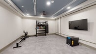 Located at 1300 Front Ave in Columbus, Georgia, this dedicated fitness room offers a thoughtfully designed space for a comprehensive workout experience. The room features a distinctive tray ceiling finished in a cool slate blue, contrasted by crisp white walls and recessed lighting that provides consistent, even illumination throughout. A ceiling fan mounted at the center of the tray ceiling ensures comfortable air circulation during intense sessions.

The flooring is a durable, smooth concrete-style surface that is both practical and easy to maintain, providing a neutral foundation that complements the room's modern aesthetic. Decorative paneling accents one wall, adding a refined architectural detail that elevates the overall design beyond a standard gym setting.

The space is equipped with a multi-functional free-standing equipment rack holding an assortment of medicine balls, kettlebells, and resistance tools, alongside a hanging heavy punching bag — ideal for strength and conditioning training. A rowing machine is positioned along one wall for cardio workouts. A wall-mounted flat-screen display enhances training sessions, while a smart interactive panel on the opposite wall adds a tech-forward element for connected fitness. The open floor plan allows ample room for stretching, bodyweight exercises, and functional movement, making this fitness room a versatile and professional-grade training environment.