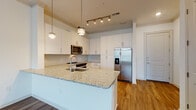 Located at 2925 Buford Dr in Buford, Georgia, this thoughtfully designed single-floor residence offers a clean, contemporary aesthetic with high-quality finishes throughout. The open-concept layout seamlessly connects the kitchen and living area, creating a bright and airy atmosphere ideal for both everyday living and entertaining.

The kitchen is a standout feature, showcasing crisp white shaker cabinetry that extends to the ceiling for ample storage, light granite countertops, and a decorative hexagonal tile backsplash. A peninsula with an undermount sink provides additional workspace and casual seating. Stainless steel appliances — including a side-by-side refrigerator with ice maker, range, microwave, and dishwasher — complete this well-appointed culinary space. Pendant lights and track lighting illuminate the area with both style and functionality.

Warm wood-style flooring runs throughout the main living areas, while bedroom spaces feature plush carpet for added comfort. The bedroom benefits from natural light through large windows with blinds, and one room includes a tray ceiling detail that adds architectural character. A balcony access door extends the living space outdoors.

The bathroom is finished with large-format wood-tone wall tiles surrounding the soaking tub and shower combination, complemented by a white vanity and wood-style flooring, delivering a spa-like feel.