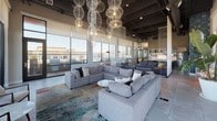 Located at 271–291 Mission Ave in Oceanside, California, this single-floor amenity space presents a thoughtfully designed communal environment that balances industrial aesthetics with contemporary comfort. The lounge area serves as the centerpiece, featuring soaring exposed ceilings with dark structural beams and an eye-catching installation of sculptural white orb pendant lights suspended at varying heights, creating a dramatic focal point above generously sized sectional sofas arranged over colorful area rugs. Floor-to-ceiling windows wrap the perimeter, flooding the interior with abundant natural light and framing panoramic outdoor views. A kitchenette and dining area anchors one end of the space, outfitted with open metal shelving, a stainless steel refrigerator, and a long communal high-top table illuminated by sleek matte black pendant lights. A bold teal patterned accent wall adds a vibrant design element. Large-format tile flooring and glass-paneled doors reinforce the modern industrial character throughout. Full-height sliding glass doors connect seamlessly to a spacious outdoor terrace furnished with lounge seating amid desert plantings, offering expansive views of the surrounding Oceanside skyline.