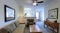 This single-floor residence showcases an open-concept layout that seamlessly connects the living, dining, and kitchen areas into one cohesive, functional space. The living area is comfortably appointed with plush seating, including a sofa and armchair arranged around a wooden coffee table, creating an inviting focal point. A ceiling fan with a built-in light fixture is centrally positioned overhead, providing both air circulation and ambient lighting. Crown molding adds a subtle decorative detail along the ceiling line.

A half-wall partition separates the kitchen from the living space while maintaining visual openness, allowing natural light from the kitchen window to flow into the main area. The kitchen features standard appliances, including a refrigerator, visible behind the partition. Beyond the living area, the dining space accommodates a table and chairs, situated near a window that draws in additional natural light. A secondary door in the dining area provides exterior access. Neutral-toned carpeting runs throughout, complementing the light-colored walls, while a wooden dresser near the entryway adds a warm, functional touch to the overall décor.