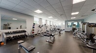 Located at 556 Colonial Rd in Franklin Lakes, New Jersey, this fitness facility offers a thoughtfully designed, single-floor space built to support a wide range of workout needs. The main exercise area is expansive and open, featuring durable rubber flooring throughout and a drop-panel ceiling with recessed fluorescent lighting that keeps the environment bright and functional. A row of windows along the perimeter wall allows natural light to flow in, providing views of the surrounding greenery.

The main gym floor is comprehensively outfitted with a robust selection of strength training machines, cable systems, free weight racks stocked with dumbbells of varying sizes, adjustable benches, and a dedicated section of cardio equipment including upright bikes, ellipticals, and treadmills. Full-length mirrors are strategically placed to support proper form during workouts.

A separate studio room, finished with warm hardwood flooring and bold teal-painted walls, provides a versatile space for stretching, personal training, or group fitness sessions. This room features wall-mounted resistance bands, balance accessories, and large mirrors. Interior windows connect the studio visually to the main gym floor. The facility is accessed through a glass-paneled entry door, with additional storage and utility rooms rounding out the overall layout.