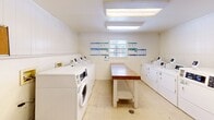 Located at 994 S Kenilworth Pkwy in Baton Rouge, Louisiana, this single-floor shared laundry facility offers a clean, efficiently organized space designed for practical use. The room is lined with white panel walls accented by crown molding, and the light tile flooring contributes to a bright, sanitary atmosphere. Overhead fluorescent lighting ensures the space is well-lit throughout.

Multiple coin-operated front-loading washers and top-loading dryers are arranged along the walls, providing ample laundry capacity. Central to the layout are two sturdy folding tables with granite-style countertops and lower shelving, offering convenient surfaces for sorting and folding laundry. A window along one wall allows in natural light and ventilation.

Informational bulletin boards are mounted on the walls, displaying usage instructions and notices for users. Safety features include a mounted fire extinguisher and visible safety signage. A dedicated payment or access control unit is also present near the entrance door, streamlining access and machine operation.

The overall layout maximizes functionality within the compact footprint, making this laundry room a well-equipped and accessible amenity for residents of the Kenilworth Parkway community in Baton Rouge.