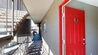 This single-floor unit makes an immediate impression with its bold red entry door, lending a vibrant, welcoming character to the exterior corridor. The building features an open-air hallway with a textured stucco exterior, offering a clean and well-maintained approach to each unit.

Inside, the spacious living area is finished with neutral wall-to-wall carpeting and soft sage-toned walls, creating a calm and inviting atmosphere. A ceiling fan with integrated lighting anchors the room, providing both comfort and practicality. The open layout connects seamlessly to the kitchen, which is equipped with dark appliances, cabinetry, and a range, all visible through a connecting passage.

A built-in shelving nook adds useful storage and architectural character to the living space, while a wall-mounted ventilation unit ensures year-round climate control. The textured ceiling adds a classic residential finish throughout the interior.

Natural light filters in through the windows, one of which frames a pleasant view of the community pool area below, surrounded by mature trees and outdoor seating. This outlook enhances the sense of openness within the unit and connects it to the broader outdoor amenities of the property.
