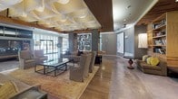 Located at 625 Monroe Street in Washington, DC, this single-floor amenity space combines contemporary design with artful sophistication. The main lounge area immediately impresses with a dramatic sculptural ceiling installation composed of layered, petal-like panels that create a dynamic overhead focal point. Seating areas are thoughtfully arranged around a large mirrored art piece and polished stone coffee tables, all set atop warm hardwood and refined stone flooring. The space transitions seamlessly into a more relaxed zone anchored by a striking fireplace with stacked wood storage, polished concrete floors, and floor-to-ceiling steel-framed glass doors that open directly to an outdoor terrace furnished for al fresco dining and lounging. A dedicated bar and kitchen area showcases reclaimed wood ceilings, a long concrete island with barstool seating, rich wood cabinetry, and an elegant inverted wine glass chandelier. Throughout the space, curated sculptural artwork and large-format wall pieces add an gallery-like ambiance. A billiards table and grand piano round out the entertainment offerings visible in the open-plan layout, while large grid-paned windows flood every area with abundant natural light. The overall design masterfully blends industrial elements with refined finishes to create a versatile, upscale communal environment.