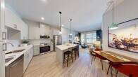 This contemporary single-floor residence offers a thoughtfully designed open-concept layout that seamlessly integrates the kitchen, dining, and living areas into one cohesive and functional space. The kitchen is a standout feature, equipped with stainless steel appliances including a French door refrigerator, range, and built-in microwave, all complemented by crisp white shaker cabinetry, quartz countertops, a textured tile backsplash, and a central island with bar seating. Pendant lighting adds a refined touch overhead, while light wood-tone flooring flows continuously throughout the residence. The living area is bright and welcoming, benefiting from large windows and a glass door that opens to a private balcony, offering views of the surrounding community. Two bedrooms provide comfortable retreats; the primary bedroom features a large window with sheer drapery, while the secondary bedroom offers a cozy, versatile layout. The bathrooms are finished with clean, modern details, including a backlit vanity mirror, white quartz countertops, shaker-style cabinetry, and wood-look tile flooring. A hallway with closet space connects the private areas of the residence. The overall ambiance is light, airy, and contemporary throughout.