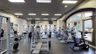 This fitness center offers a comprehensive workout environment designed to accommodate a wide variety of exercise routines. The space features a generous open floor plan with a dark rubber flooring that provides both durability and traction throughout the workout area. Overhead, recessed panel lighting ensures the entire space is brightly and evenly illuminated, complemented by natural light streaming in through multiple large windows along the perimeter walls, creating an energizing and welcoming atmosphere. The ceiling is finished in a neutral tone and outfitted with ceiling fans, contributing to air circulation and comfort during workouts. The facility is stocked with an impressive array of equipment, including treadmills, elliptical machines, upright stationary bikes, and cable weight machines, as well as a free weights section complete with a barbell rack and bench press station. Multiple flat-screen televisions are mounted at eye level throughout the space, allowing members to stay entertained or informed during their sessions. An arched architectural detail on one wall adds a refined design touch, elevating the overall aesthetic beyond a standard gym environment. The combination of thoughtful layout, ample equipment variety, and quality finishes makes this fitness center a well-rounded and functional space for all fitness levels.