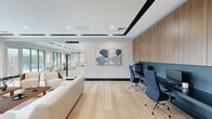 This contemporary single-floor space offers a thoughtfully designed blend of collaborative lounge and functional workspace areas. Light hardwood flooring runs throughout, complementing a clean, neutral color palette of whites and soft grays accented by bold black trim details on the ceiling coffers and baseboards.

The main lounge area is anchored by a generous seating arrangement featuring cream leather sofas and modern walnut-toned accent chairs arranged around low lattice-style coffee tables, creating an inviting setting for meetings or relaxed gatherings. Large, multi-pane windows line the exterior walls, flooding the interior with natural light and offering open views of the surrounding greenery and urban streetscape.

A dedicated workstation zone is integrated along one wall, featuring a teal-backed built-in desk fitted with ergonomic task chairs and warm wood cabinetry above, providing a practical yet polished workspace. Abstract artwork adorns the walls, adding a refined artistic touch.

Floor-to-ceiling glass partitions with sleek black metal frames divide the space, maintaining visual openness while defining distinct functional zones. Recessed ceiling lighting and subtle tray ceiling detailing further elevate the overall aesthetic, contributing to a professional yet welcoming ambiance throughout.