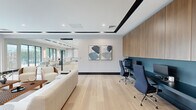 This contemporary single-floor space offers a thoughtfully designed blend of collaborative lounge and functional workspace areas. Light hardwood flooring runs throughout, complementing a clean, neutral color palette of whites and soft grays accented by bold black trim details on the ceiling coffers and baseboards.

The main lounge area is anchored by a generous seating arrangement featuring cream leather sofas and modern walnut-toned accent chairs arranged around low lattice-style coffee tables, creating an inviting setting for meetings or relaxed gatherings. Large, multi-pane windows line the exterior walls, flooding the interior with natural light and offering open views of the surrounding greenery and urban streetscape.

A dedicated workstation zone is integrated along one wall, featuring a teal-backed built-in desk fitted with ergonomic task chairs and warm wood cabinetry above, providing a practical yet polished workspace. Abstract artwork adorns the walls, adding a refined artistic touch.

Floor-to-ceiling glass partitions with sleek black metal frames divide the space, maintaining visual openness while defining distinct functional zones. Recessed ceiling lighting and subtle tray ceiling detailing further elevate the overall aesthetic, contributing to a professional yet welcoming ambiance throughout.