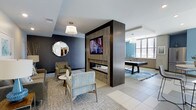 Located at 28 Woolfe St in Charleston, South Carolina, this single-floor community lounge space offers a thoughtfully designed environment that blends comfort and entertainment. The interior is anchored by a striking dark wood accent wall that serves as both a room divider and a focal point, housing a linear gas fireplace below a wall-mounted flat-screen television. Adjacent to the lounge seating area, a decorative geometric mirror adds visual interest to the deep charcoal-toned feature wall. The lounge area is furnished with modern striped armchairs and a small side table, all set atop a patterned area rug over large-format tile flooring, creating a warm and inviting atmosphere. Flowing seamlessly from the lounge is a game room area, highlighted by a sleek billiards table centered on a graphic area rug. This space benefits from generous natural light streaming through expansive windows, which also frame views of the surrounding neighborhood. A wet bar or kitchenette with cabinetry lines the far wall, providing convenient amenity access. The color palette of blues, grays, and warm wood tones throughout gives the space a cohesive, contemporary aesthetic well-suited for social gatherings.