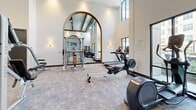 This fitness center is designed with both functionality and aesthetics in mind, offering a spacious, single-floor layout beneath dramatically high ceilings that create an open, airy atmosphere. The architecture incorporates a distinctive arched mirror feature with warm wood trim, serving as a visual centerpiece that divides the space while enhancing depth and light throughout the room.

Large floor-to-ceiling windows and black-framed glass panels line the walls, flooding the interior with natural light and providing views of an outdoor pool area and surrounding greenery. The neutral-toned carpet flooring and crisp white walls contribute to a clean, modern aesthetic.

The cardio section is well-equipped with multiple elliptical machines, rowing machines, and a stair climber, all positioned to take advantage of the exterior views. A dedicated strength training area features a cable machine, a multi-function weight rack, free weights including a full dumbbell rack, and a weight bench. Built-in open shelving along one wall provides organized storage for accessories. A wall-mounted flat-screen television is positioned for easy viewing during workouts. The entrance features dark-framed double glass doors, completing the polished, upscale feel of this comprehensive fitness facility.