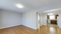 This single-floor space presents a straightforward and functional layout with a clean, minimalist aesthetic. The main room is finished with light-toned hardwood flooring that runs throughout, contributing warmth and continuity to the overall design. Neutral gray-white walls keep the interior feeling bright and open, complemented by white baseboards that provide a crisp contrast to the flooring.

A flush-mount ceiling light fixture provides overhead illumination, ensuring the space remains well-lit. The smooth, flat ceiling maintains a simple, uncluttered look. An open doorway connects this main room to an adjacent area, where natural light is visible, suggesting a window in the neighboring space. This passthrough opening enhances the sense of flow between rooms.

Further beyond the doorway, additional rooms are partially visible, hinting at a connected layout that includes what appears to be a kitchen or utility area with darker wood cabinetry. A baseboard heater is also visible along one wall, indicating a practical heating solution integrated into the space. Overall, the interior offers a clean, move-in-ready environment with consistent finishes and a neutral palette suitable for a variety of uses.