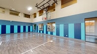 This single-floor indoor sports facility is centered around a full-sized basketball court finished with a durable hardwood-style floor featuring clearly marked regulation court lines. The space is defined by impressively high ceilings that accommodate a wall-mounted, glass-backboard basketball hoop with a retractable mechanism overhead. The perimeter walls are finished in a two-tone blue color scheme—deep navy on the upper half and bright teal padding panels on the lower half—providing both aesthetic cohesion and player safety. Safety padding lines the walls consistently around the court, reflecting a well-considered design for active use. Large floor-to-ceiling windows along one side of the court flood the space with natural light while offering views of the surrounding outdoor landscape, including mature trees. Elevated interior windows are also visible near the ceiling on one wall, suggesting an adjacent observation or mezzanine area. Seating areas with metal chairs are positioned along the sidelines, accommodating spectators. Glass double doors provide access to an adjacent fitness area visible through the panes. Recessed and track lighting supplements natural light throughout the court.