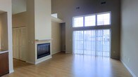 This two-floor space is defined by its dramatic vertical proportions and open, airy design. The main living area immediately draws attention with its soaring double-height ceilings, which amplify the sense of space and allow natural light to flood the interior through a generous bank of large upper windows and expansive floor-to-ceiling sliding glass doors. These doors open to an outdoor area and are dressed with vertical blinds, offering both privacy and light control.

The living area features smooth, light-toned hardwood flooring that extends throughout, contributing to a clean and cohesive aesthetic. A built-in fireplace with a sleek white mantel serves as a focal point along one wall, adding warmth and architectural character to the space. The surrounding walls are finished in a neutral warm beige tone, complementing the minimalist design.

The towering chimney column anchors the room while highlighting the vertical scale of the interior. HVAC vents are integrated discreetly into the upper walls and ceilings. The overall ambiance is modern, open, and bright, offering a versatile layout well-suited for comfortable living.