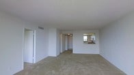 This single-floor space presents an open and functional layout centered around a generously sized living area. The room is finished with neutral-toned flooring and white walls, creating a clean, understated aesthetic throughout. A textured ceiling adds subtle visual interest overhead, while a ceiling vent ensures consistent climate control.

A standout architectural detail is the pass-through window connecting the kitchen to the main living area, allowing for convenient interaction between the two spaces. The kitchen, visible through the opening, features overhead cabinetry and benefits from natural light through a window, lending it a bright and practical character.

A hallway extends from the living area, leading to additional rooms within the unit, including what appears to be a bedroom or secondary space visible in the background. Doors along the hallway suggest private, separated areas within the layout.

The overall ambiance is bright and open, with a straightforward design that provides a versatile canvas for personalization. The minimal detailing and neutral palette make the space adaptable to a wide range of furnishing styles and functional needs.