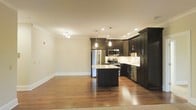 This single-floor space showcases a thoughtfully designed open-concept layout that seamlessly connects the kitchen and living areas. The kitchen is a standout feature, highlighted by rich, dark-toned cabinetry that extends to the ceiling, offering generous storage. Light-toned granite countertops provide a striking contrast against the dark cabinetry, while a central island with pendant lighting above serves as both a functional workspace and a visual focal point. Stainless steel appliances, including a refrigerator, range, microwave, and dishwasher, lend a modern, polished feel to the space. A white subway tile backsplash adds texture and brightness to the cooking area. Warm hardwood flooring runs throughout, enhancing the inviting ambiance of the space. Recessed lighting, complemented by a multi-bulb track fixture, ensures the area is well-lit. Crown molding along the ceiling adds an architectural detail that elevates the overall finish. The open flow between the kitchen and the adjacent living area creates a spacious, functional environment well-suited for both everyday living and entertaining.