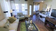 This single-floor residence showcases an open-concept layout that effortlessly connects the living, dining, and kitchen areas into one cohesive, flowing space. Rich, dark hardwood flooring runs throughout, providing a warm and elegant foundation that anchors the entire interior.

The living area is bathed in natural light, courtesy of large windows dressed with floor-length white curtains accented by bold dark trim, lending a crisp, tailored look. The space is well-proportioned, comfortably accommodating a plush sofa, a patterned accent chair, and a glass-top coffee table adorned with decorative candle centerpieces and a vibrant green area rug that adds a lively pop of color.

Transitioning seamlessly from the living area, the dining space features a dark wood dining table and chairs positioned near additional windows, ensuring the area remains bright and airy. A breakfast bar with tall bar stools provides a casual alternative dining option and serves as a natural divider between the kitchen and the rest of the open-plan space.

The kitchen is clean and functional, with light countertops that contrast beautifully against the darker tones of the flooring and cabinetry. Throughout the space, the interplay of natural light, warm wood tones, and thoughtful décor accents creates an inviting and comfortable ambiance ideal for both everyday living and entertaining.