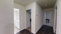 This single-floor residence offers a clean, contemporary aesthetic defined by neutral tones and quality finishes throughout. The interior is anchored by rich, dark hardwood flooring that runs through the main hallway and living areas, providing a striking contrast against the crisp white walls and trim. The hallway serves as the central artery of the home, connecting multiple rooms with an open and airy flow. A well-designed bathroom is accessible directly from the corridor, featuring white cabinetry with ample storage, a vanity with countertop space, and classic tile flooring with a decorative pattern that adds a subtle design detail. The bedroom, visible through an adjoining doorway, continues the dark hardwood flooring and benefits from the home's overall sense of calm and light. White door frames and baseboards provide clean architectural definition throughout, while the neutral wall palette keeps the space feeling bright and versatile. The layout is practical and efficient, with each room thoughtfully positioned for ease of access. Overall, this residence presents a polished, move-in-ready environment with consistent design choices that create a cohesive and inviting atmosphere from room to room.