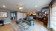 This single-floor residence offers a cohesive, modern aesthetic anchored by a cool blue-grey color palette and warm wood-tone flooring that flows seamlessly throughout the space. The open-concept living area serves as the heart of the home, featuring a ceiling fan, recessed lighting, and a comfortable sectional sofa arranged around a glass-topped coffee table. The space transitions naturally into a dedicated dining area and a fully equipped kitchen. The kitchen is outfitted with stainless steel appliances including a gas range and refrigerator, two-tone cabinetry featuring dark lower cabinets and white uppers, a tile backsplash, and a peninsula that opens toward the living space. Two bedrooms offer distinct personalities: the primary bedroom is bathed in natural light from multiple windows with custom window treatments, and features a ceiling fan and a closet, while the second bedroom showcases a reclaimed wood headboard with industrial accents and carpeted flooring. Both bathrooms are finished with wood-look tile flooring, white vanities, and sliding glass-door shower enclosures. Throughout the residence, thoughtful décor choices, including framed artwork and decorative mirrors, contribute to a polished and inviting atmosphere.