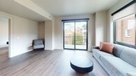 This well-designed 2-bedroom, 2-bathroom apartment offers 800 square feet of thoughtfully laid-out single-floor living space. The open kitchen is a standout feature, equipped with stainless steel appliances including a refrigerator, range, microwave, and dishwasher. Two-tone cabinetry in soft gray and beige is complemented by a white subway tile backsplash, white countertops, open wooden shelving, and a built-in dining nook — creating a functional and stylish culinary space. The living room is bright and airy, highlighted by large floor-to-ceiling windows and a sliding glass door that opens to a private balcony with views of the surrounding neighborhood. Light hardwood-style flooring flows consistently throughout the unit. Both bedrooms are well-proportioned, each featuring large windows with roller shades that allow for natural light and privacy. The primary bedroom measures 150 square feet. Both bathrooms follow a clean, neutral aesthetic with light cabinetry, white countertops, tub/shower combinations with curved curtain rods, and hardwood-style flooring. The overall design of the unit is modern and cohesive, emphasizing natural light, clean lines, and a neutral color palette throughout.