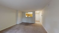 This single-floor unit features a practical, well-organized layout connecting the living areas, kitchen, bedroom, and bathroom in a cohesive flow. The spacious living room is finished with wall-to-wall carpeting in a neutral tone and features a pass-through window opening directly into the kitchen, promoting an open, connected feel. A sliding glass door leads to a private balcony overlooking lush greenery, filling the room with natural light. A wall-mounted heating unit adds a functional element to the living space.

The kitchen is efficiently designed with warm oak cabinetry, dark granite-style countertops, and tile flooring. White appliances, including an electric range, dishwasher, and over-the-range microwave, are neatly integrated. A stainless steel sink is positioned at the corner of the L-shaped counter layout.

The bedroom is generously sized with plush carpeting, a window with natural light, and a wall-mounted air conditioning unit. The full bathroom features a tiled tub-surround, a toilet, tile flooring, and a wall-mounted cabinet providing additional storage. Throughout the unit, neutral walls and clean finishes create a bright, understated ambiance.